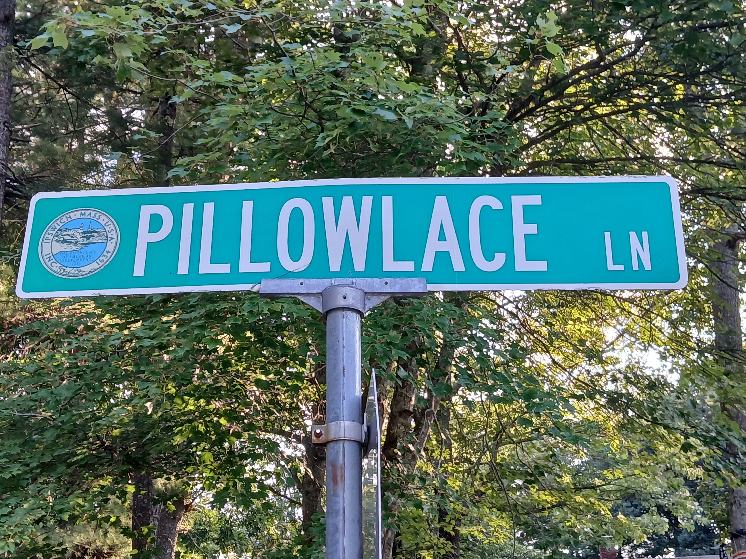 A city street sign in Ipswich MA that has the city seal on the left and the text PILLOWLACE LN