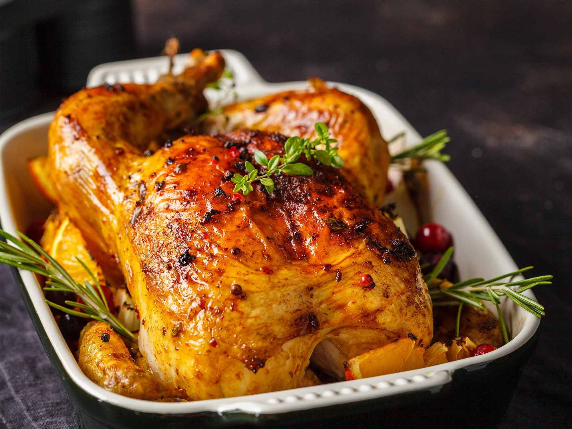 Roast chicken garnished with herbs in a white baking dish with roasted vegetables and cranberries.