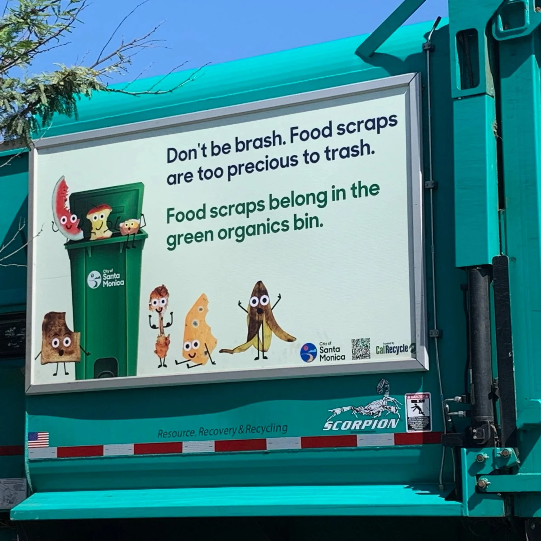 Sign promoting food scraps recycling with animated food characters around a green trash bin, mentioning food scraps belong in the green bin in Santa Monica.