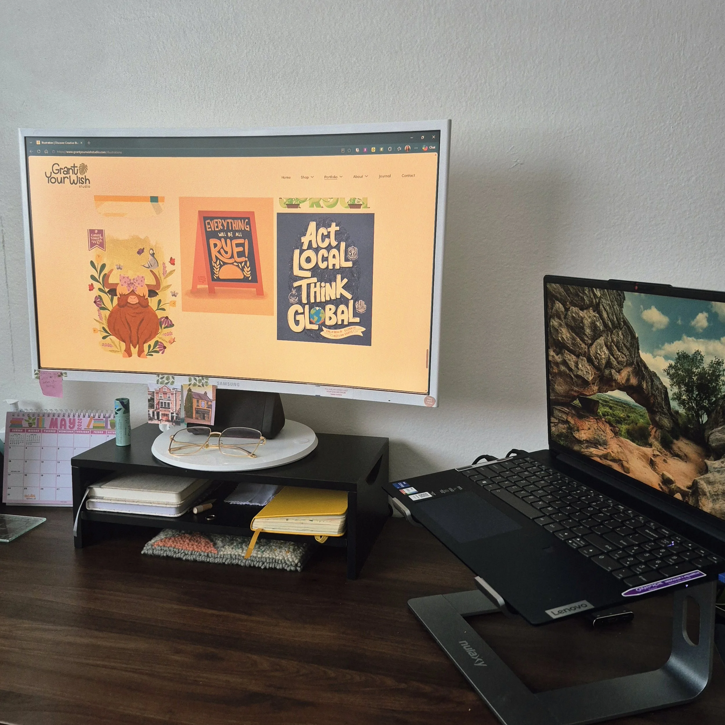 A workspace with a computer monitor displaying a colorful website, a calendar, a small notebook, and glasses on a dark wooden desk. Another monitor is partially visible on the right side.