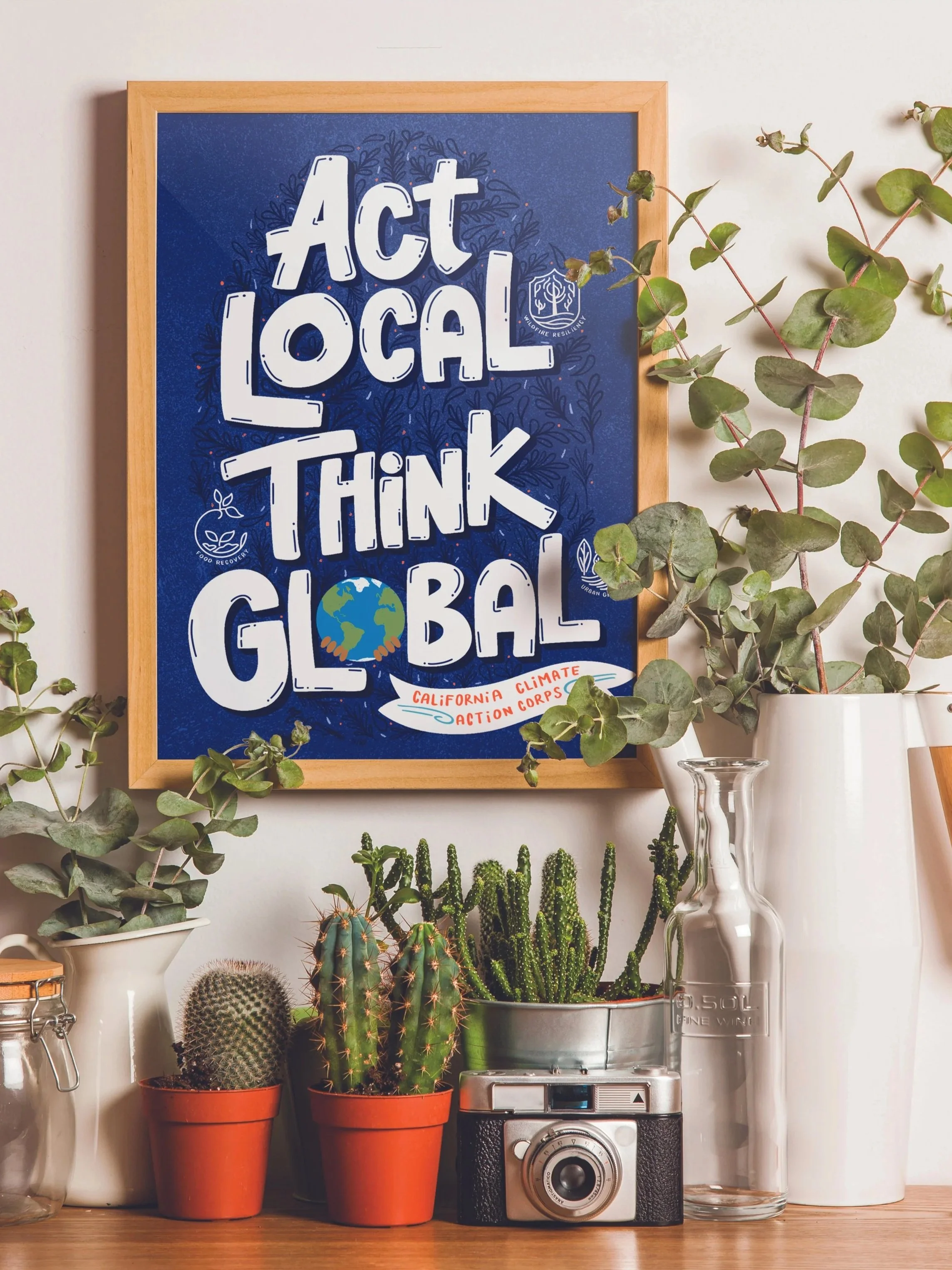 A table with various potted cacti, green plants, a vintage camera, and glass vases. A framed poster with a blue background reads 'Act Local Think Global' with logos and a globe illustration.