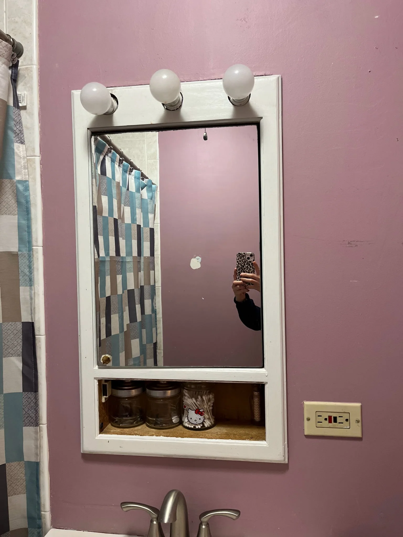 Bathroom mirror with three round light bulbs above it, purple wall, wooden shelf with jars, and a medicine cabinet area before renovation