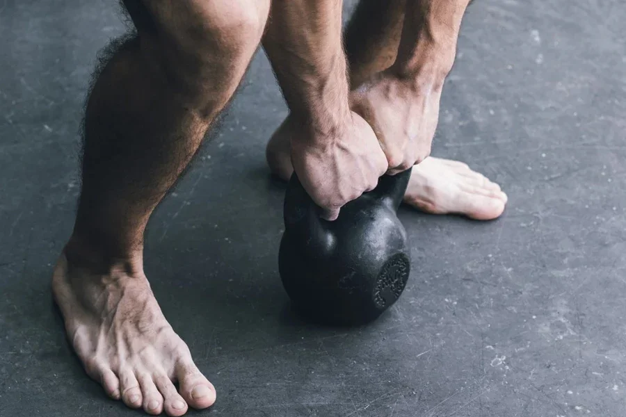 A person is performing a strength exercise with a black kettlebell, standing barefoot on a gym floor.