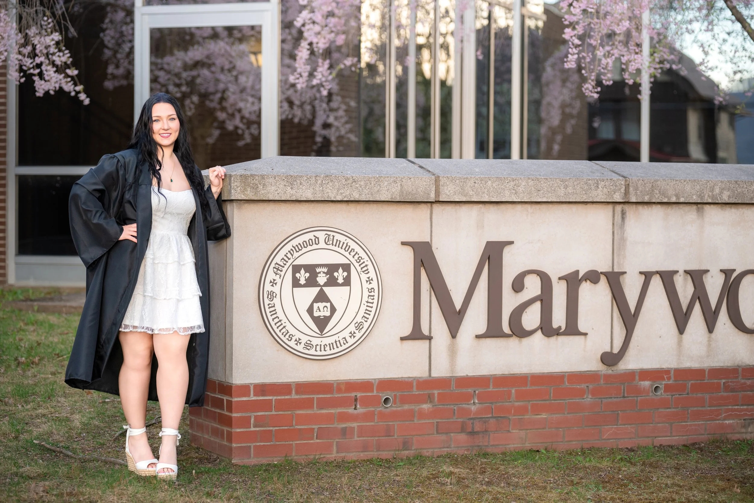 A young woman in a white dress and black graduation gown posing next to a stone sign that says "Marywood" with the university's emblem next to it. She is smiling and standing outdoors with pink flowering trees reflected in the windows behind her.