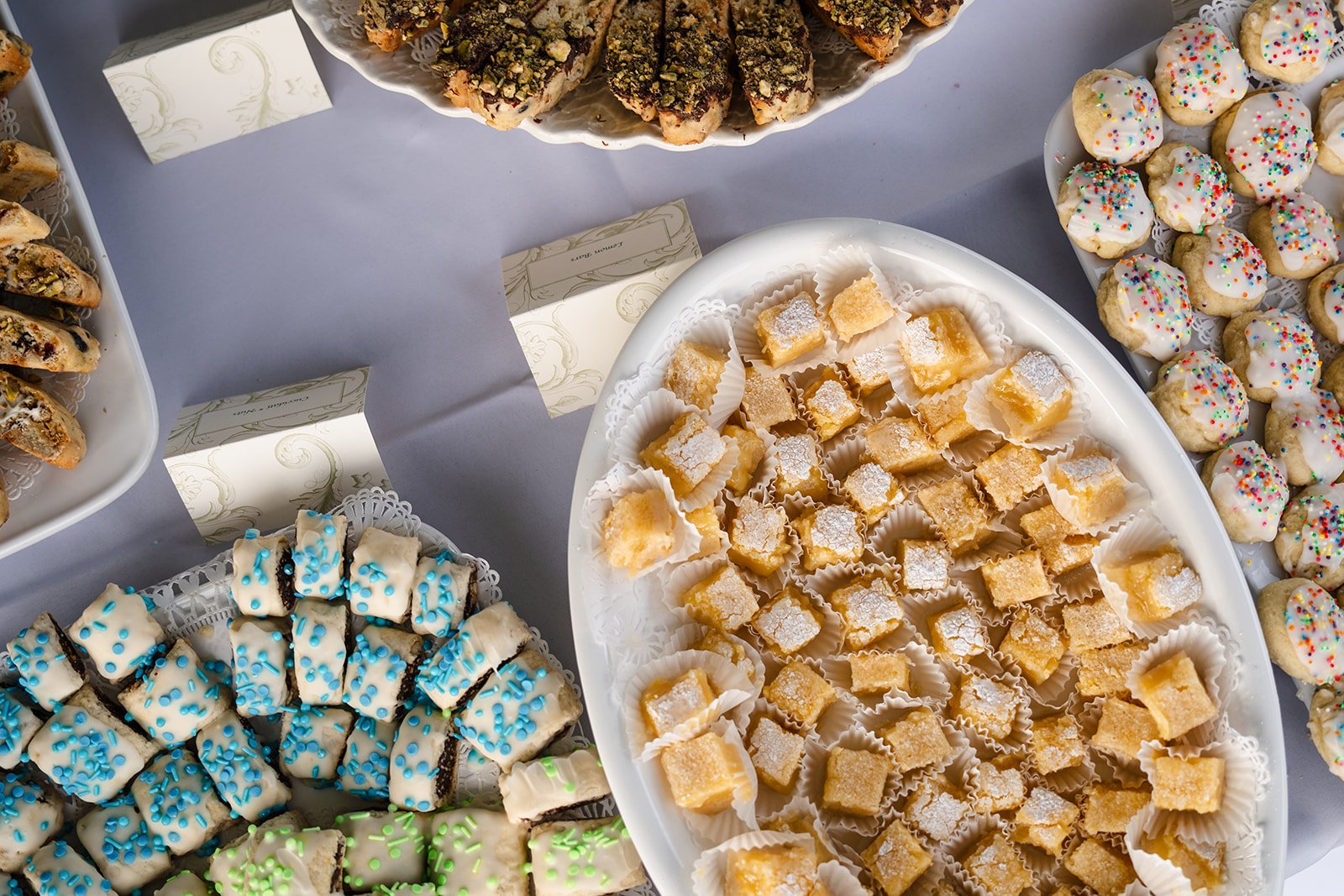 Assorted cookies and pastries on platters, including biscotti, lemon bars, and iced cookies with sprinkles.