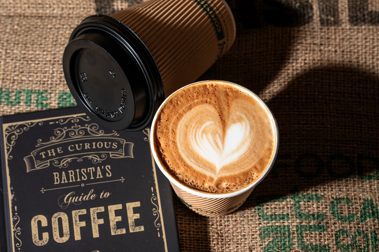 A cup of latte with heart-shaped latte art next to a lid and a book titled "The Curious Barista's Guide to Coffee" on burlap.