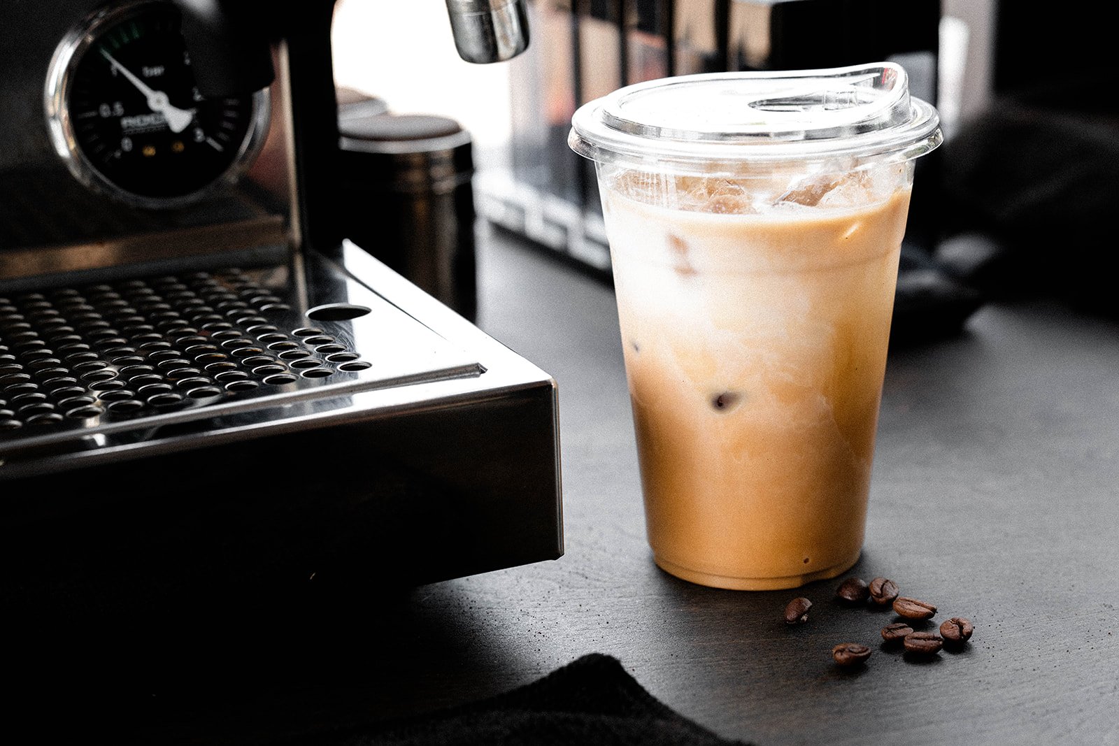 Iced coffee in a plastic cup next to an espresso machine and coffee beans on a table.