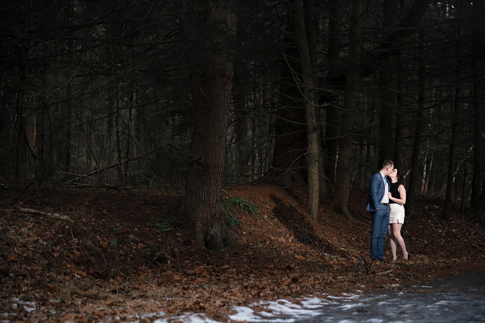 An engaged couple kissing in a forest, standing on a path with fallen leaves and snow.