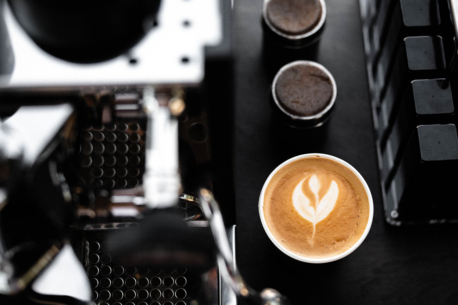 A cup of coffee with latte art next to an espresso machine and two portafilter baskets on a counter.