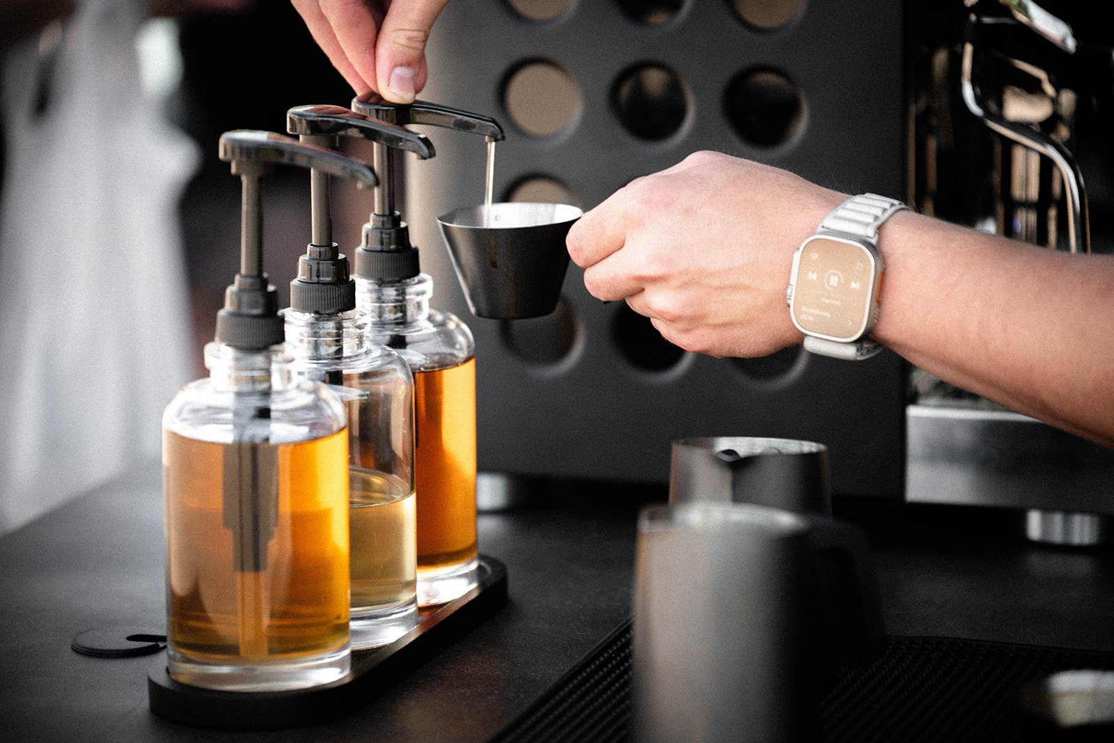 Person dispensing syrup from pump bottles into a small cup, with a watch on their wrist.