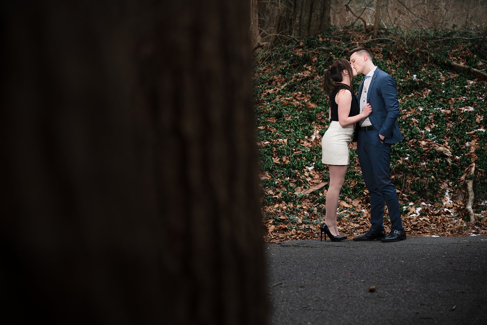 Engaged couple kissing near a tree in a forested area with fallen leaves and greenery.