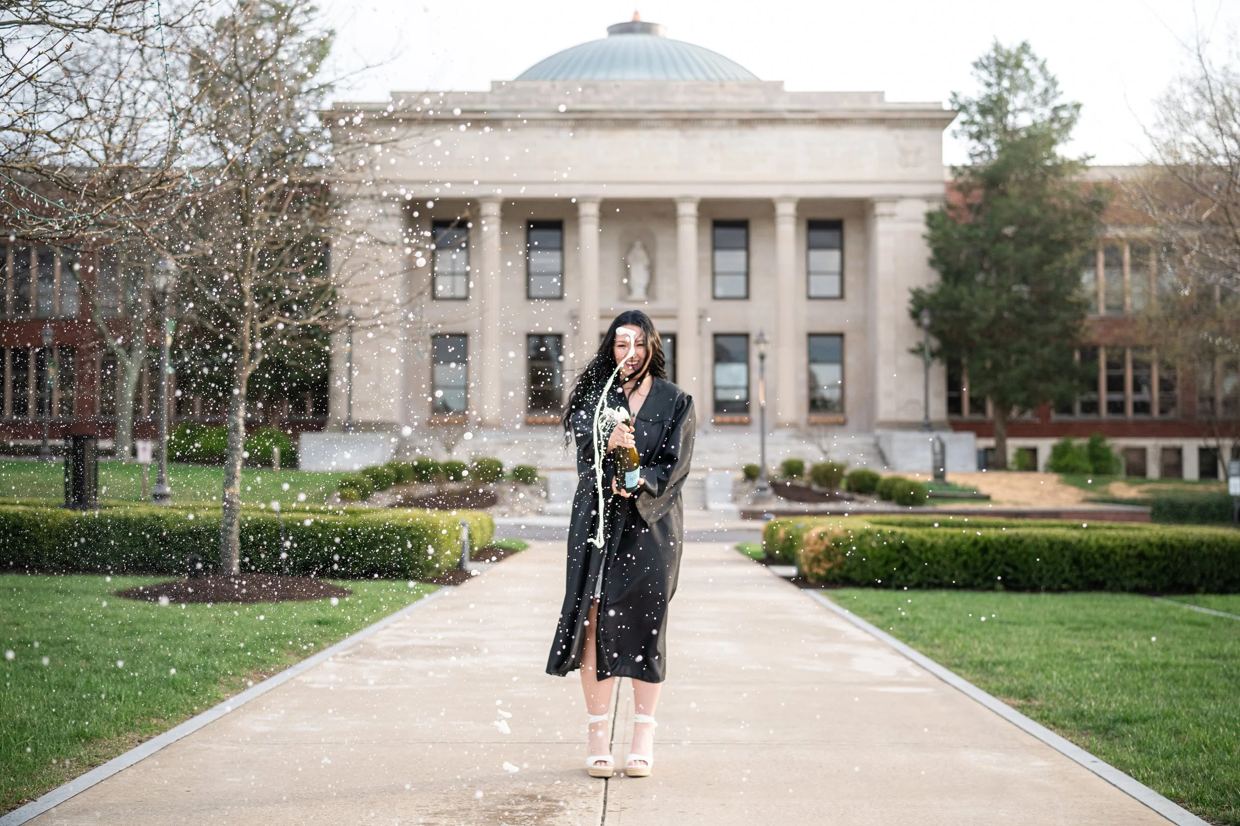 A woman in a graduation gown celebrating with a champagne bottle on a university campus, with a large historic building in the background and confetti in the air.