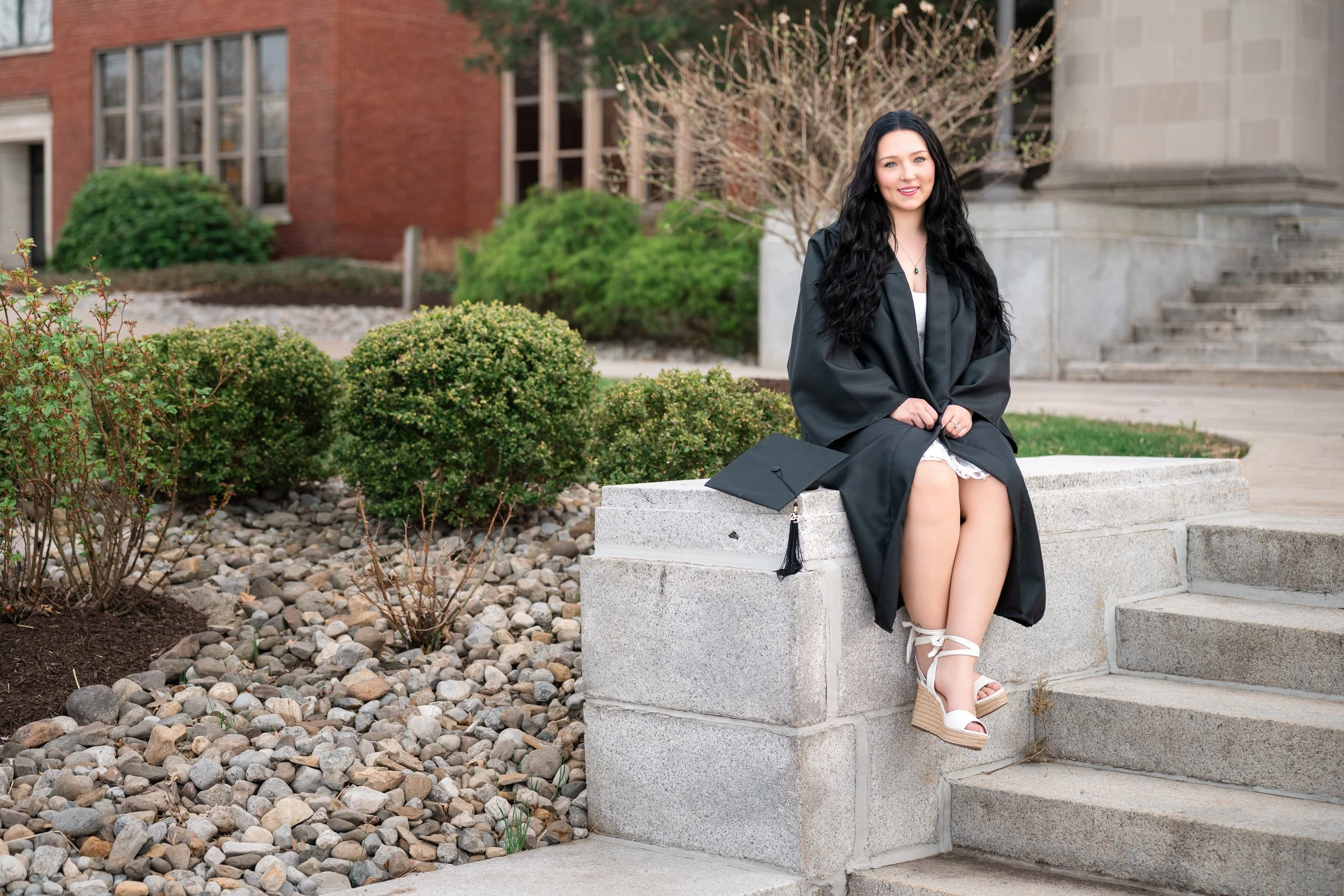 A young woman in a black graduation gown sitting on a concrete ledge outdoors with a graduation cap placed beside her. She is smiling and wearing white wedge sandals, with steps and a garden with bushes and trees in the background.