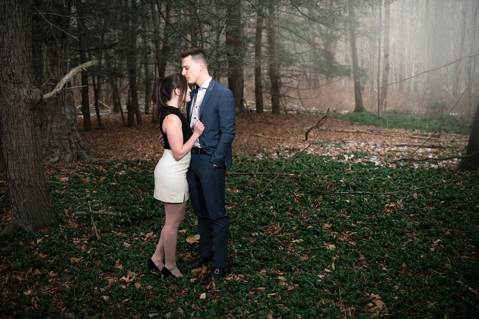 An engaged couple in formal attire standing closely in a forest, with fallen leaves and green ground cover, surrounded by tall trees.