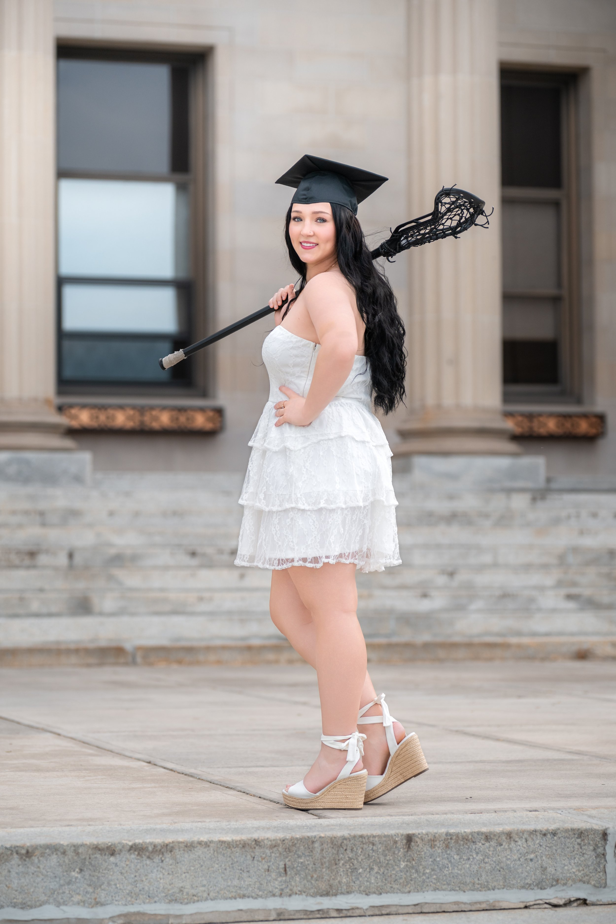 Young woman in a white dress with a graduation cap holding a lacrosse stick, standing outside on steps in front of a building.