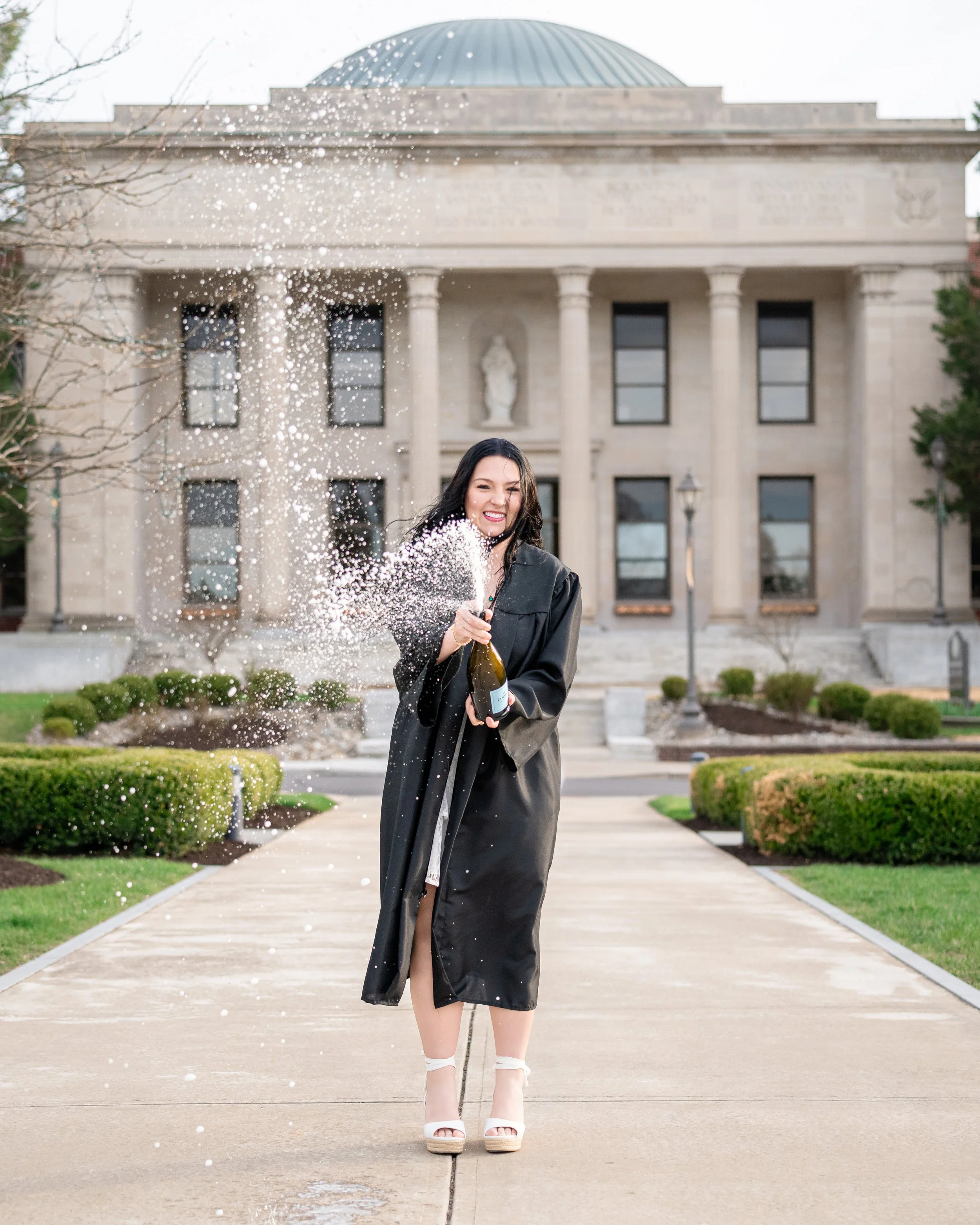 A woman in a graduation gown celebrating outside a university building with a bottle of champagne, popping the cork, causing champagne and foam to fly into the air.