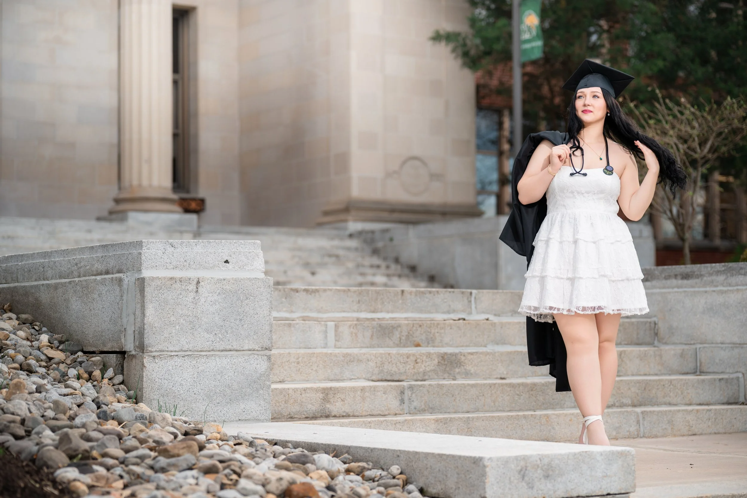 A young woman in a white dress holding a black graduation gown over her shoulder, wearing a graduation cap and standing on stairs outside a building.