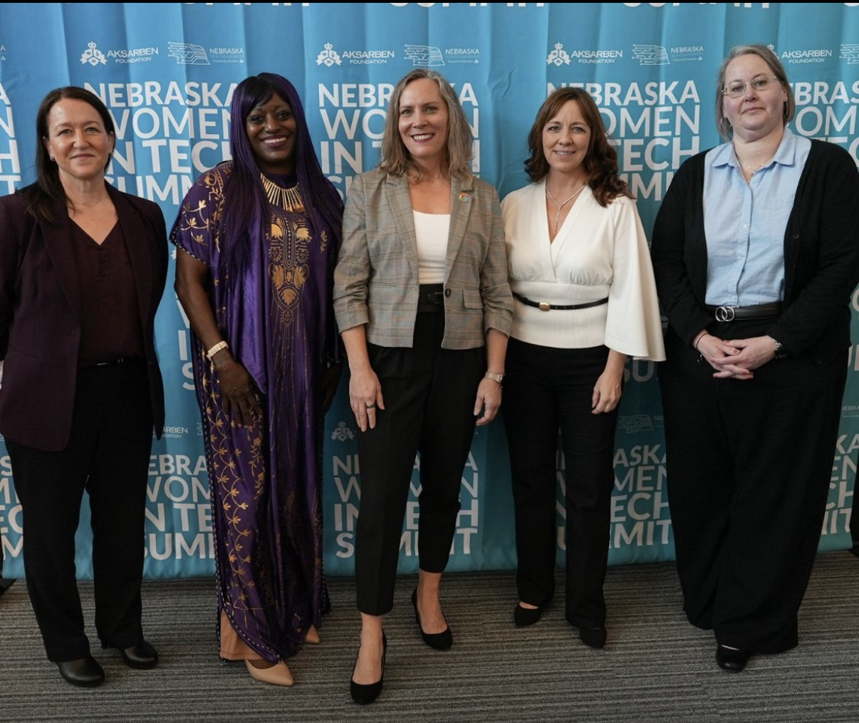 Five women standing side by side in front of a blue Nebraska Women in Tech Summit backdrop.