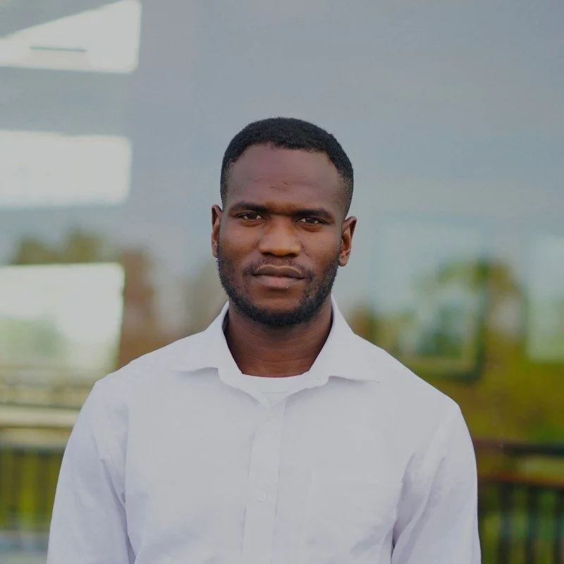 A young man with dark hair and a beard, wearing a white shirt, standing outdoors in front of a glass building with reflections of trees and sky.