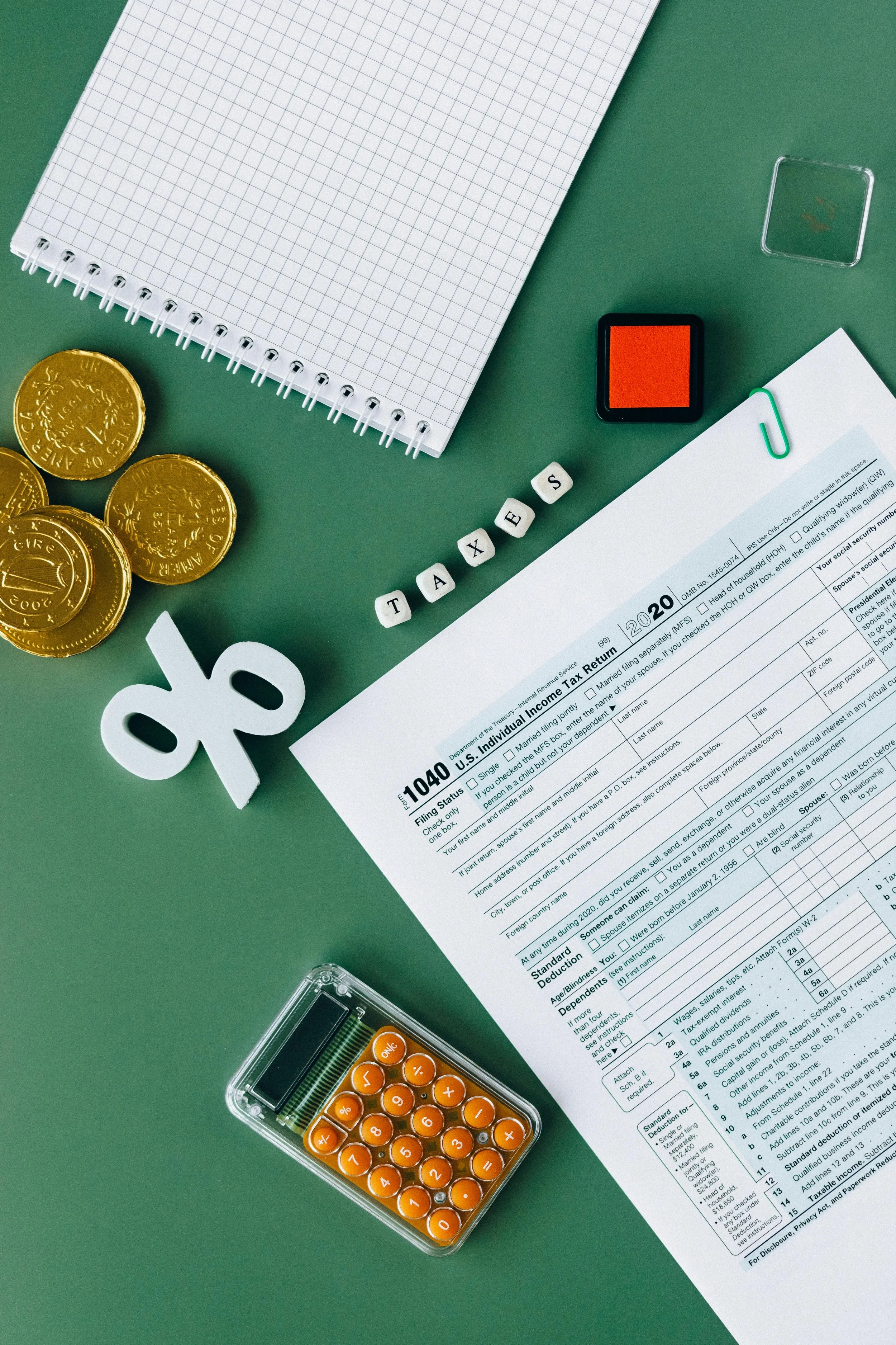 A green desk with a 2020 U.S. individual tax return form, a notebook with graph paper, a small stamp pad, a paperclip, a few gold coins, a white key-shaped symbol, a miniature calculator, and letter beads spelling 'TAXES'.