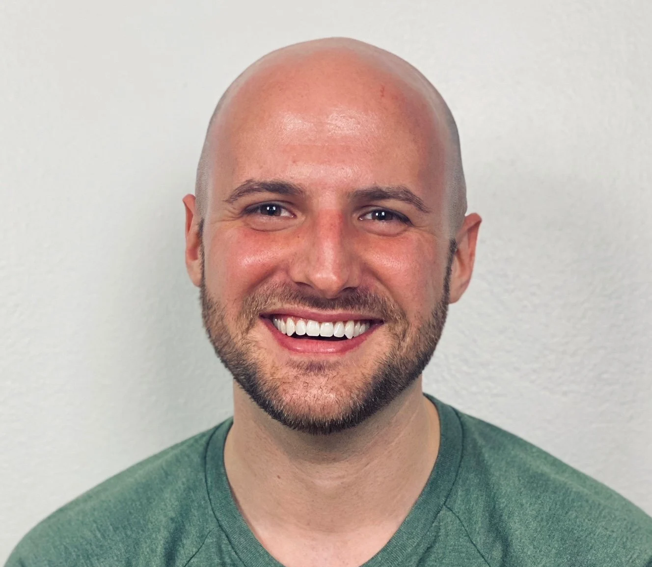 Smiling man with a shaved head, beard, and mustache, wearing a green t-shirt, standing against a plain white wall.