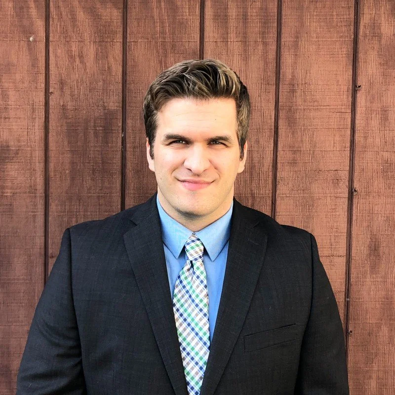 A young man in a dark suit and a colorful plaid tie, standing in front of a wooden wall.