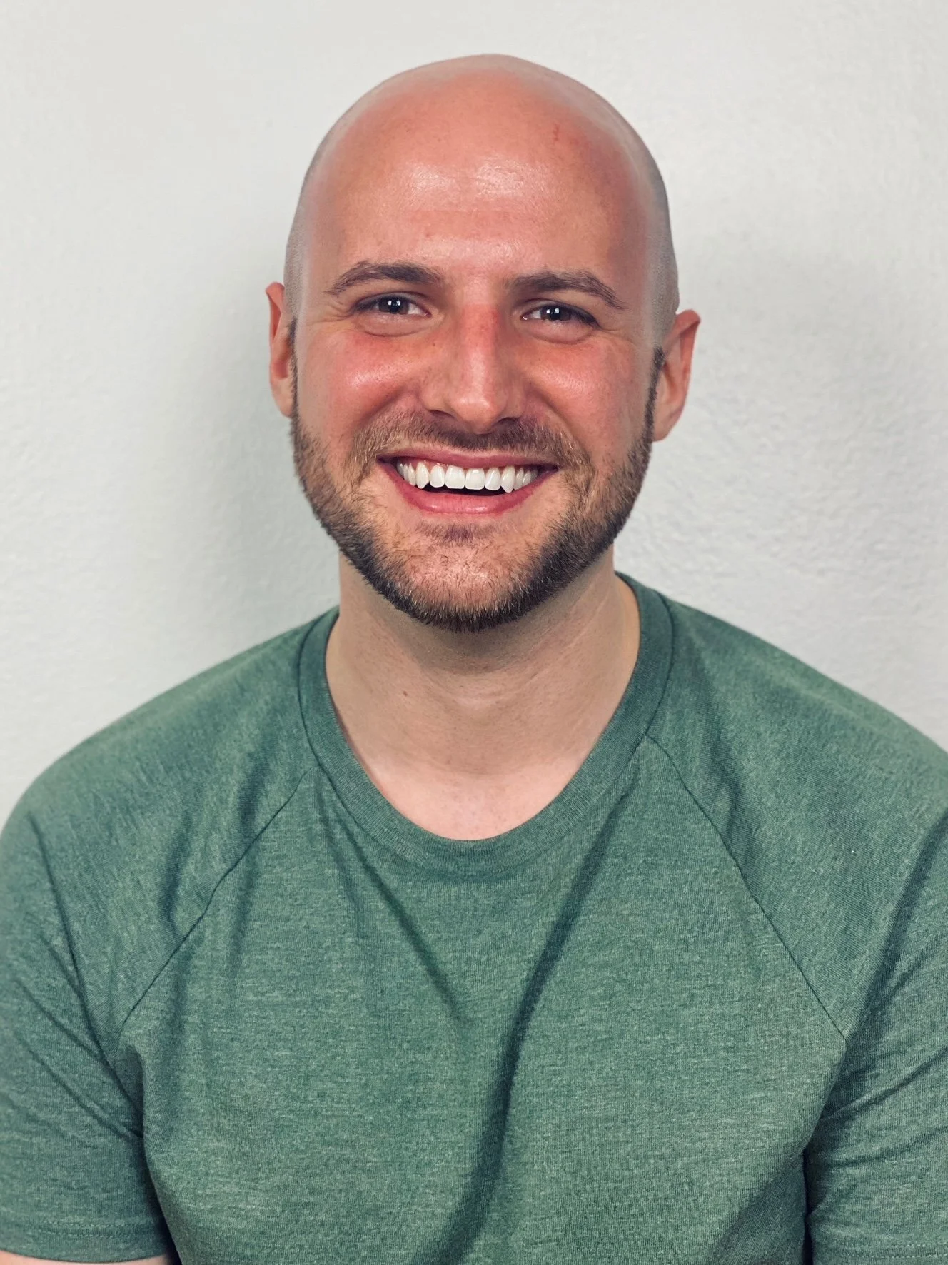 Smiling man with a shaved head, beard, and mustache, wearing a green t-shirt, standing against a plain white wall.