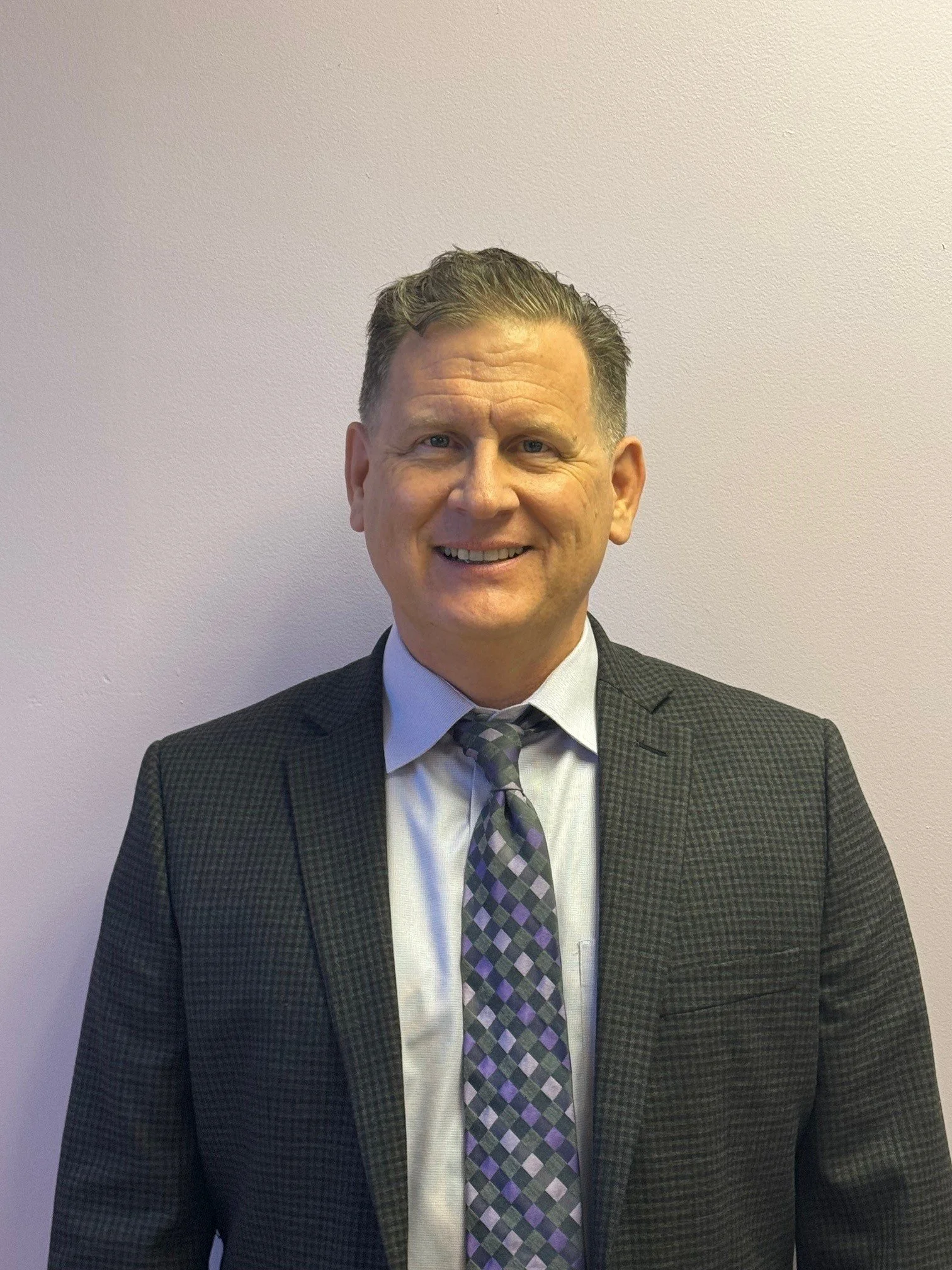 A middle-aged man wearing a black checkered suit jacket, a light blue dress shirt, and a multicolored checkered tie, standing against a plain beige wall and smiling at the camera.