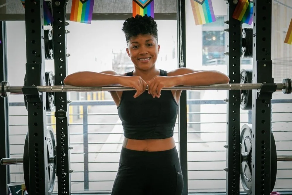 Marci standing at the squat bar smiling with rainbow pride flags above.
