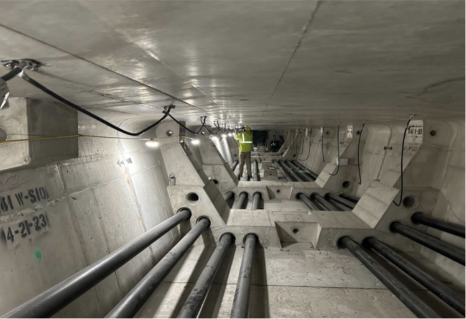 Construction worker in safety vest working inside a deep underground utility or maintenance shaft with large pipes and concrete walls.