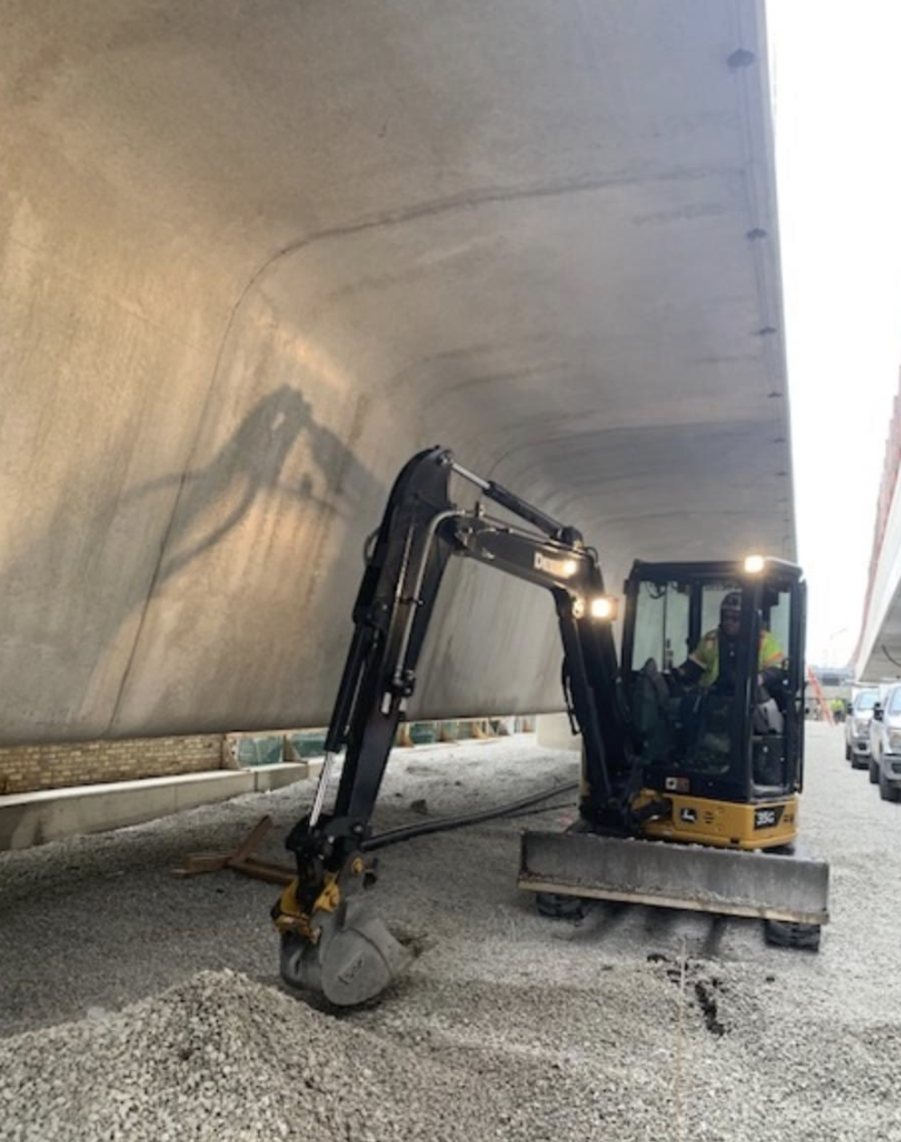 A construction worker operating a small excavator under a large concrete bridge or structure. The worker is wearing a hard hat and safety vest.