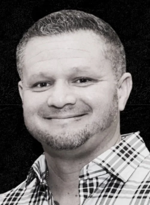Black and white photo of a smiling man with short hair, a goatee, wearing a checkered shirt with a dark background.