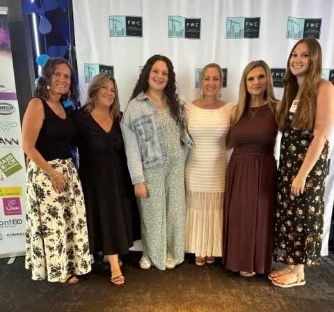 Six women standing in front of a white backdrop with logos, smiling for the photo, at an indoor event.