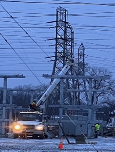 A utility worker in a yellow vest repairing high-voltage power lines on a utility pole using an aerial lift truck, with electrical infrastructure and trees in the background.