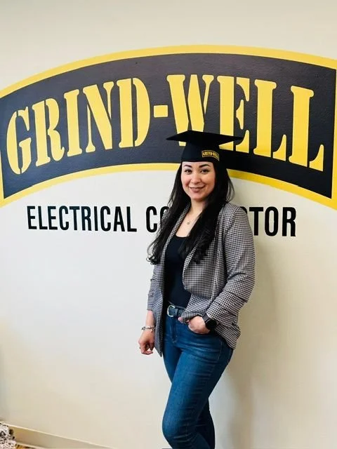 Woman in graduation cap and casual outfit standing in front of a wall with a sign that reads 'GRIND-WELL ELECTRICAL CONTRACTOR'