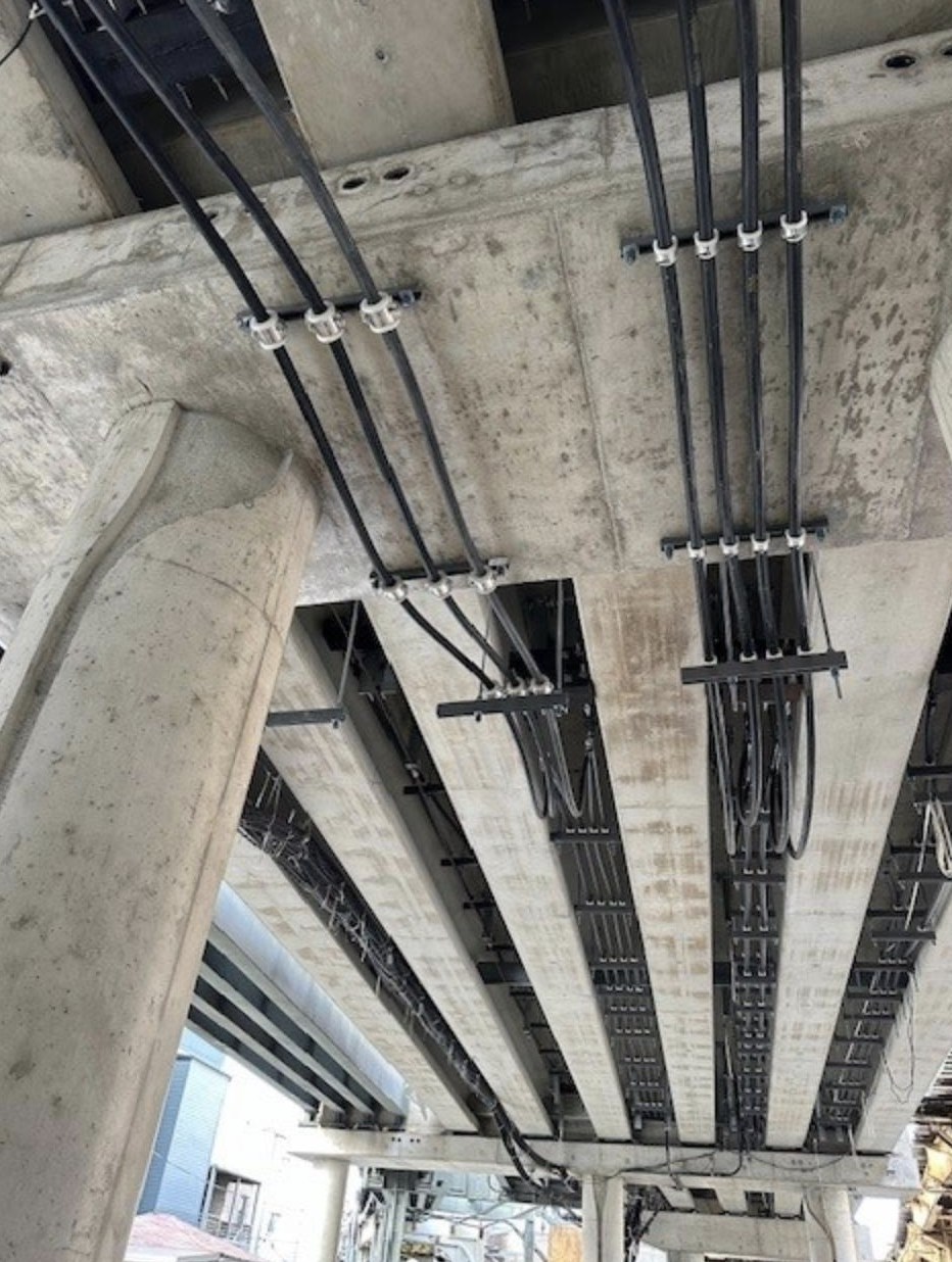 View of electrical wiring and conduit pipes installed on the underside of a concrete structure at a construction site.