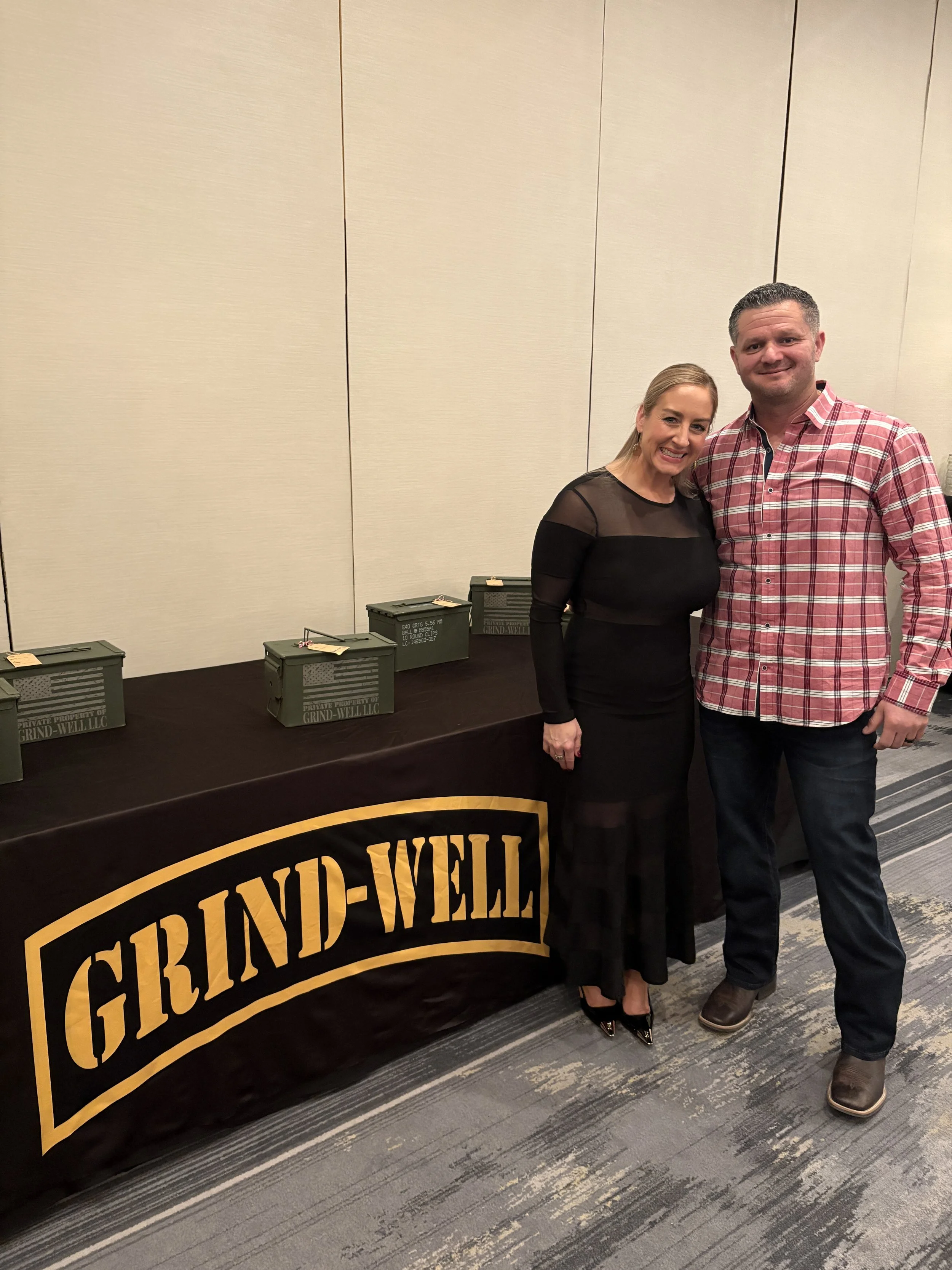 A man and woman are standing side by side, smiling at the camera, in front of a table with boxes on it. The table has a black cloth with a large yellow and black sign that reads 'GRIND-WELL LLC'. The woman is wearing a black dress with sheer sleeves,
