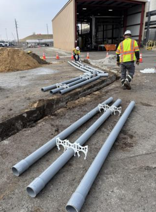 Construction workers installing large gray pipes on the ground outside a building under construction, with orange traffic cones and a dirt pile nearby.