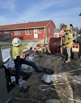 Two firefighters with protective gear and helmets work with a large red hose on a trailer, with a red wooden house and blue sky in the background.