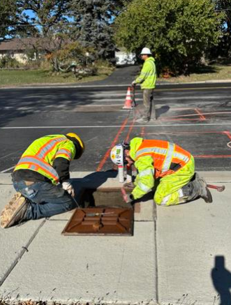 Two construction workers in high-visibility safety vests and helmets kneeling on a sidewalk, marking lines for road work with orange spray paint, while another worker stands further back with traffic cones.