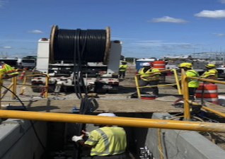 Construction workers and equipment on a construction site, with orange and white safety barrels, yellow guard rails, and a large spool of cable on a truck, under a clear blue sky.
