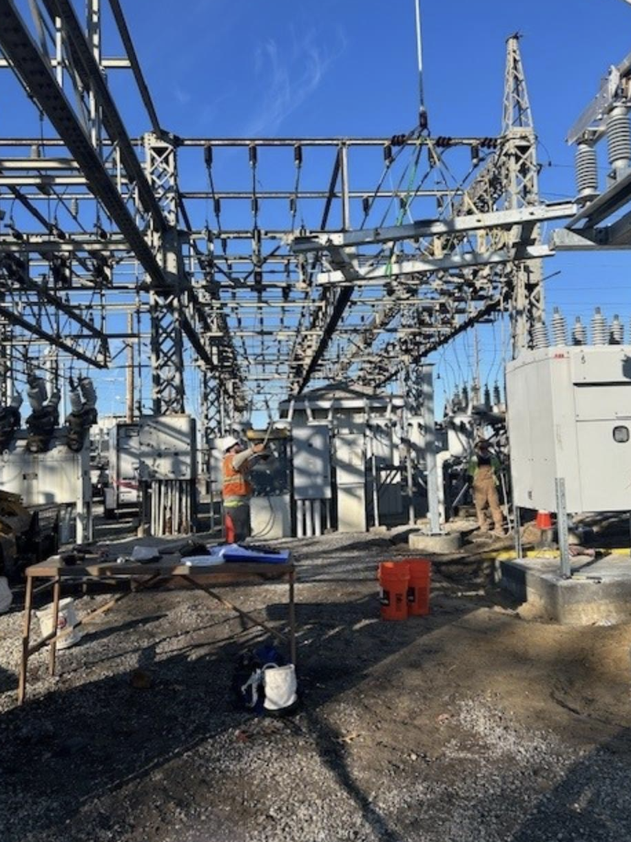 Workers at a power substation performing maintenance or construction work under a blue sky.