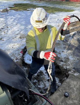 Worker in safety gear holding a rope with a hook at the end, outdoors in a snowy area, near an open utility access point.