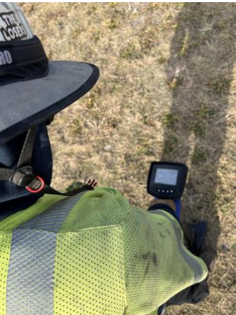 Person wearing a yellow safety vest and a black hat, using a metal detector outdoors on grassy terrain.