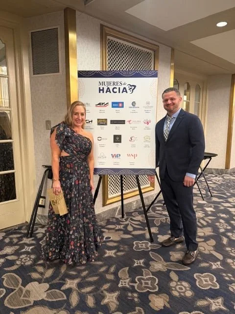 A woman and a man dressed in formal attire standing next to a large event sign at the Mujeres de Hacia event, with a patterned carpet and decorative wall paneling in the background.