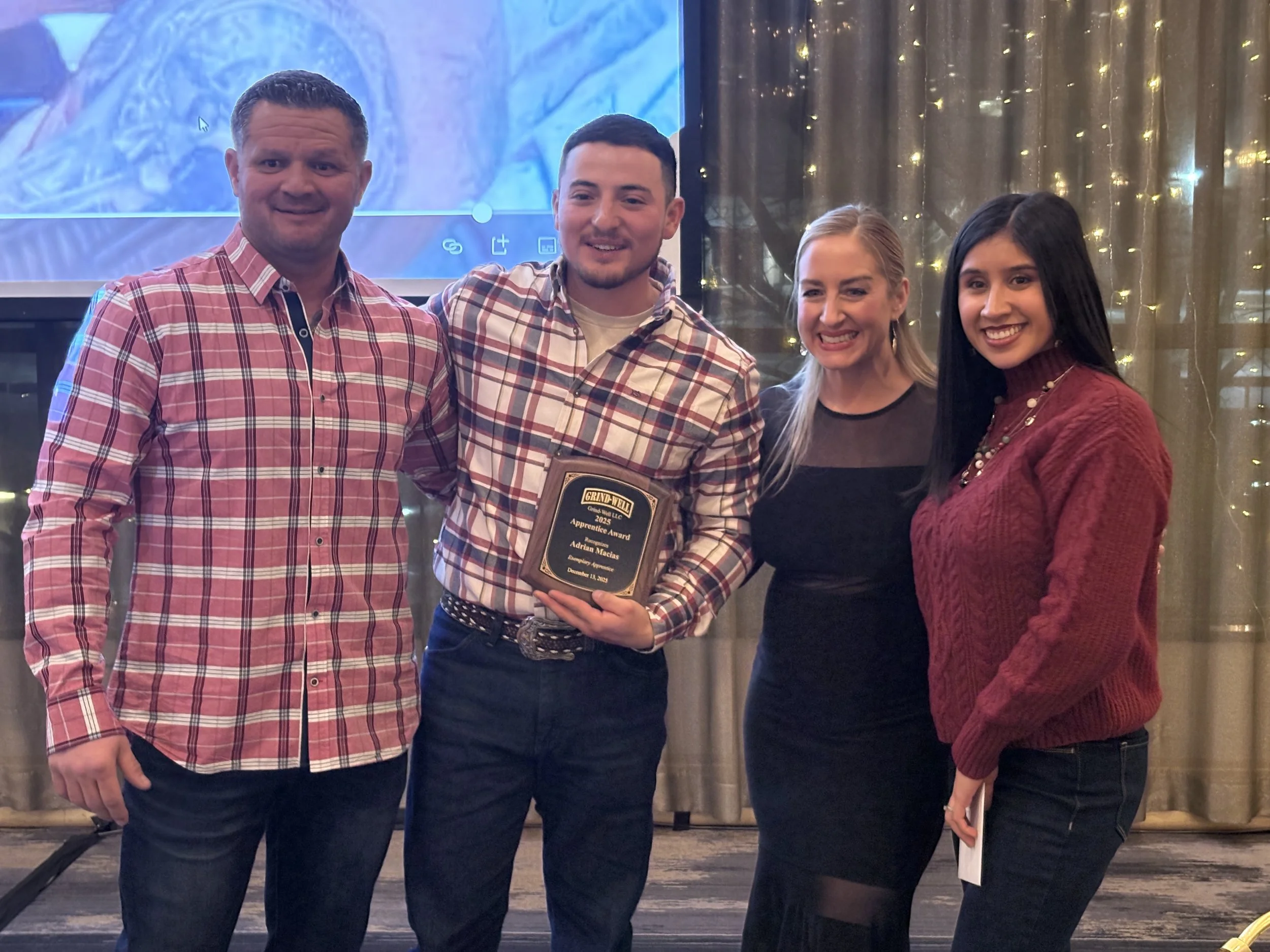 Four people standing together at an event. The second person from the left is holding an award plaque with the text 'Grand-Well' and 'Apprentice Award 2023'. All are smiling, dressed casually, with a background of string lights and a large screen.