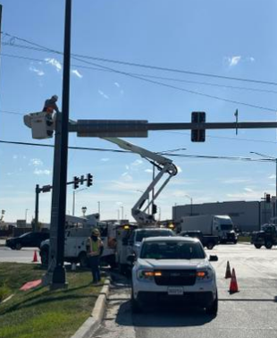 Utility worker in a bucket lift repairing traffic lights at an intersection, with cars and traffic cones nearby.