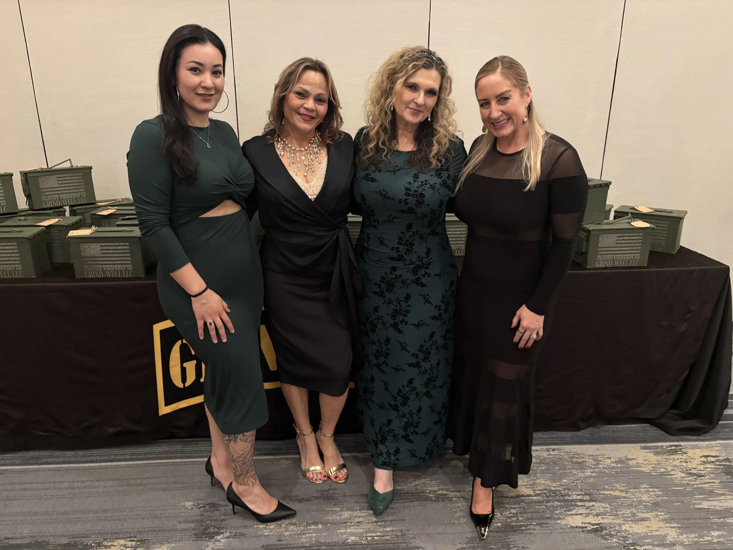 Four women standing side by side in front of a table with green boxes, smiling at the camera, dressed in evening attire.