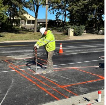 Worker in a neon yellow safety vest marking lines on a city street with spray paint and orange cones.