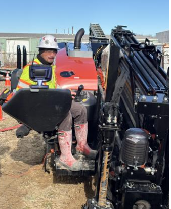 A construction worker wearing a safety helmet, reflective vest, and rubber boots operating a small orange construction vehicle at a work site.